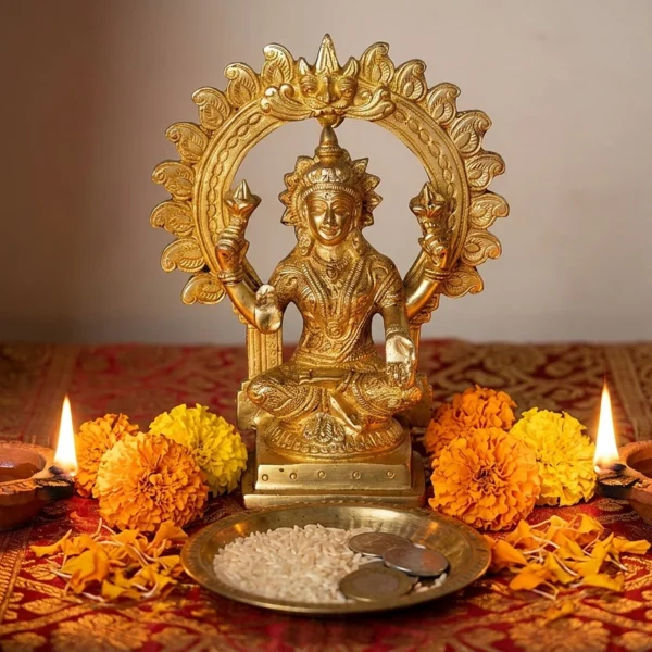 Brass Lakshmi idol with carved arch, 7 inch, on a red and gold cloth with lit diyas, marigolds and a plate of rice and coins for Diwali puja